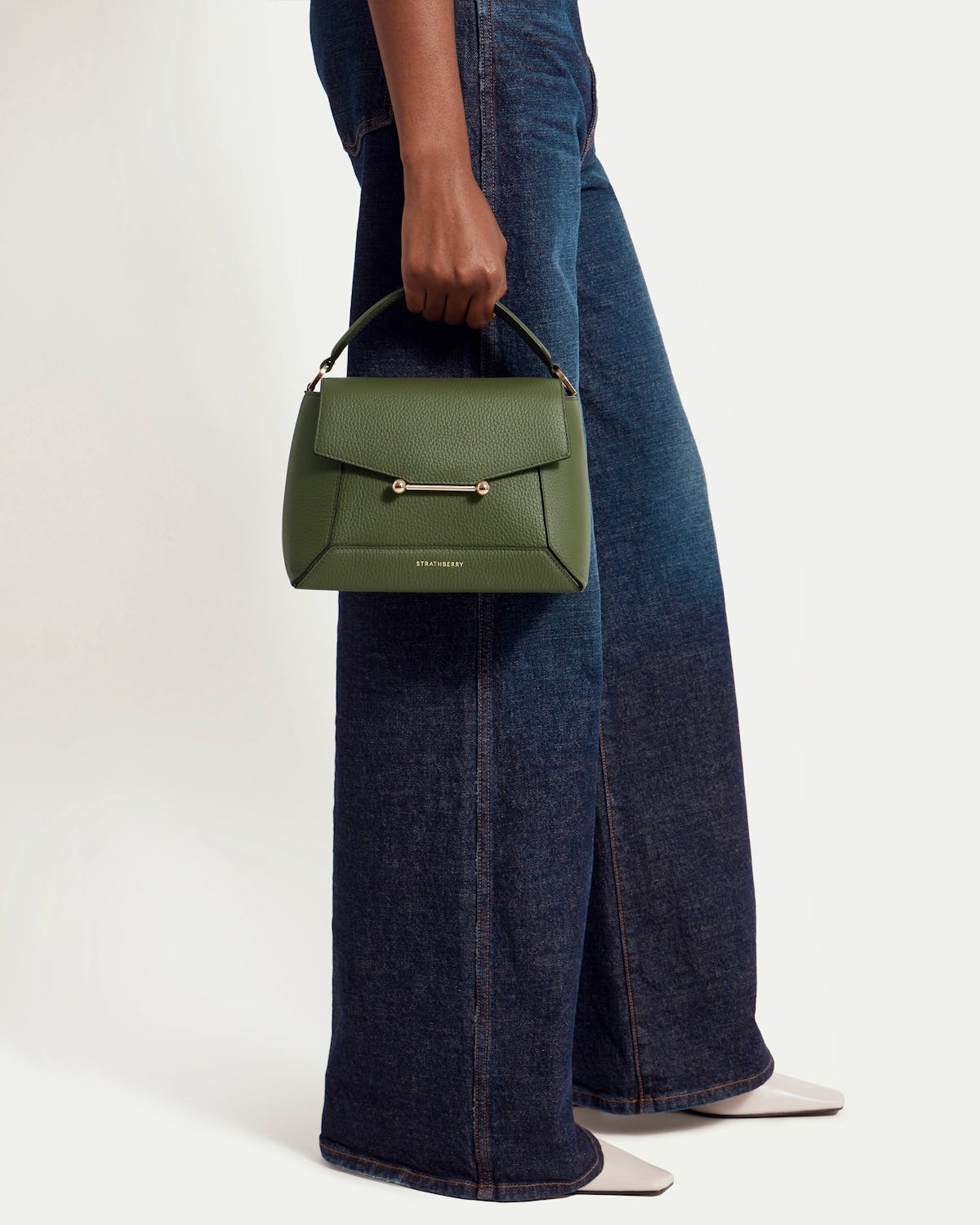 Model wearing dark wide-leg jeans holds a pine green Strathberry Mosaic bag.