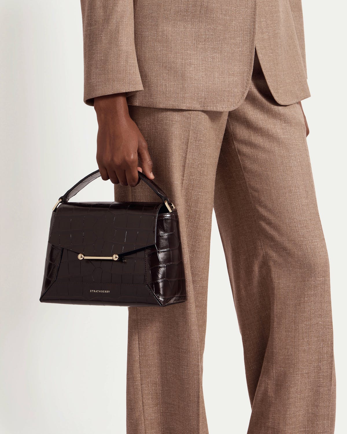 Model holding croc-embossed mosaic bag with top handle and Strathberry logo, wearing brown suit.