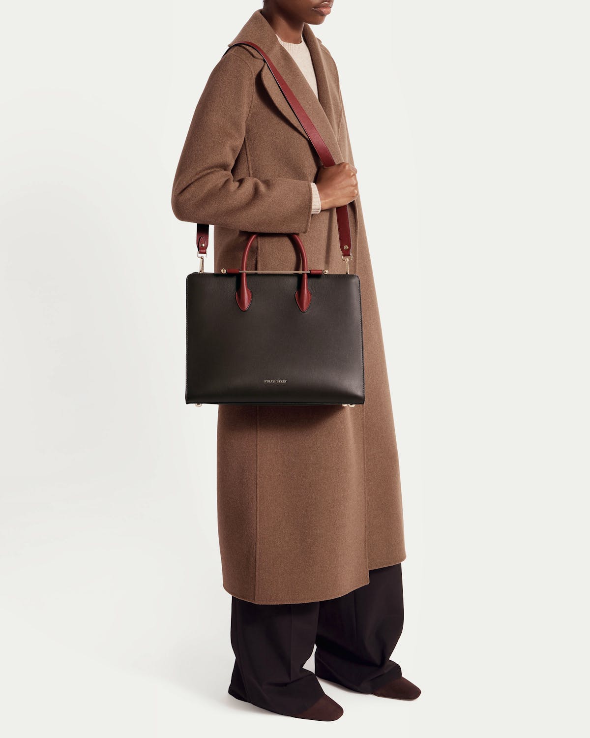 Woman in brown coat holding Strathberry espresso tote with oxblood handles and strap.