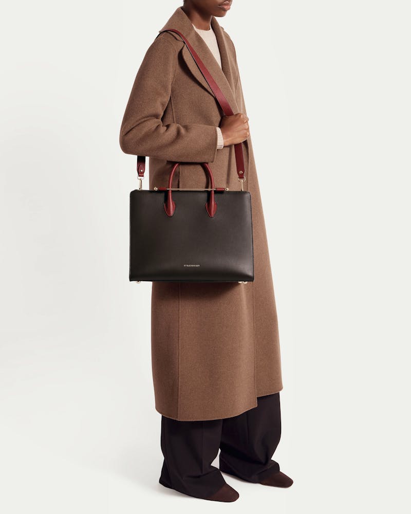 Woman in brown coat holding Strathberry espresso tote with oxblood handles and strap.