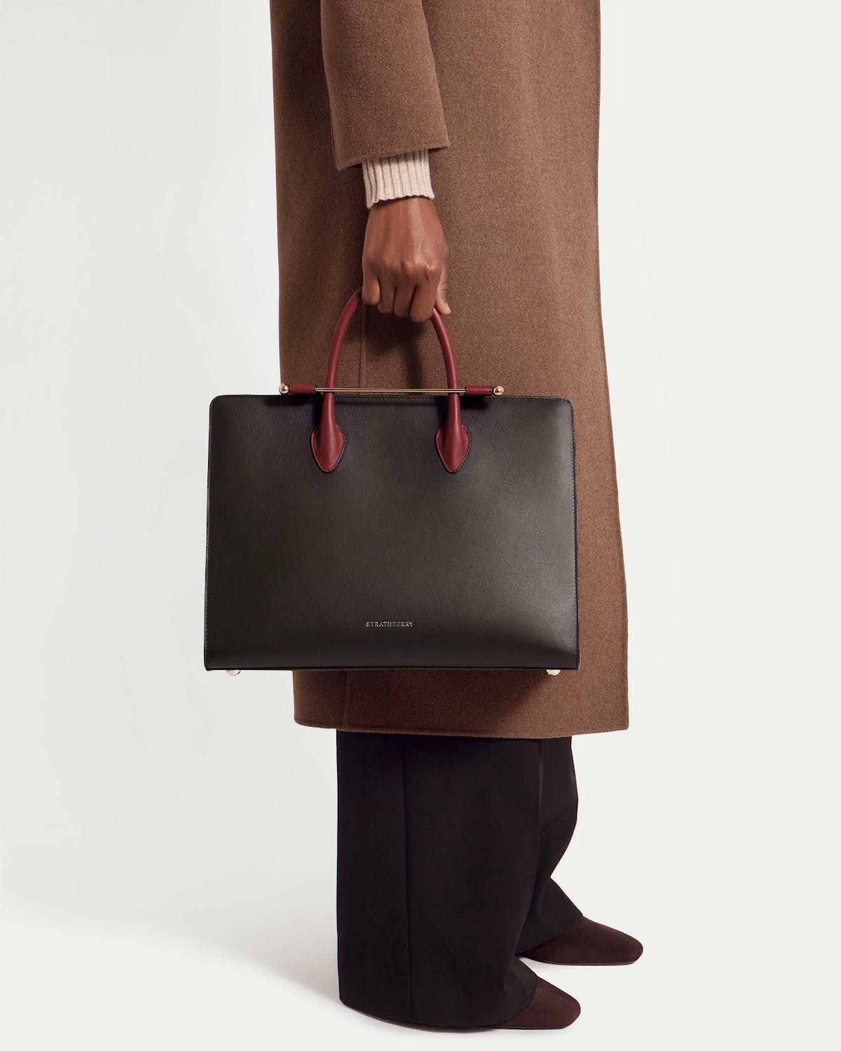 Model holding a Strathberry tote with espresso body, oxblood handles and black trim.