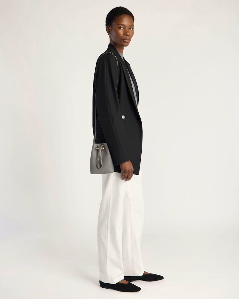 Model in black blazer and white wide-leg trousers carrying stone grey midi pouch.