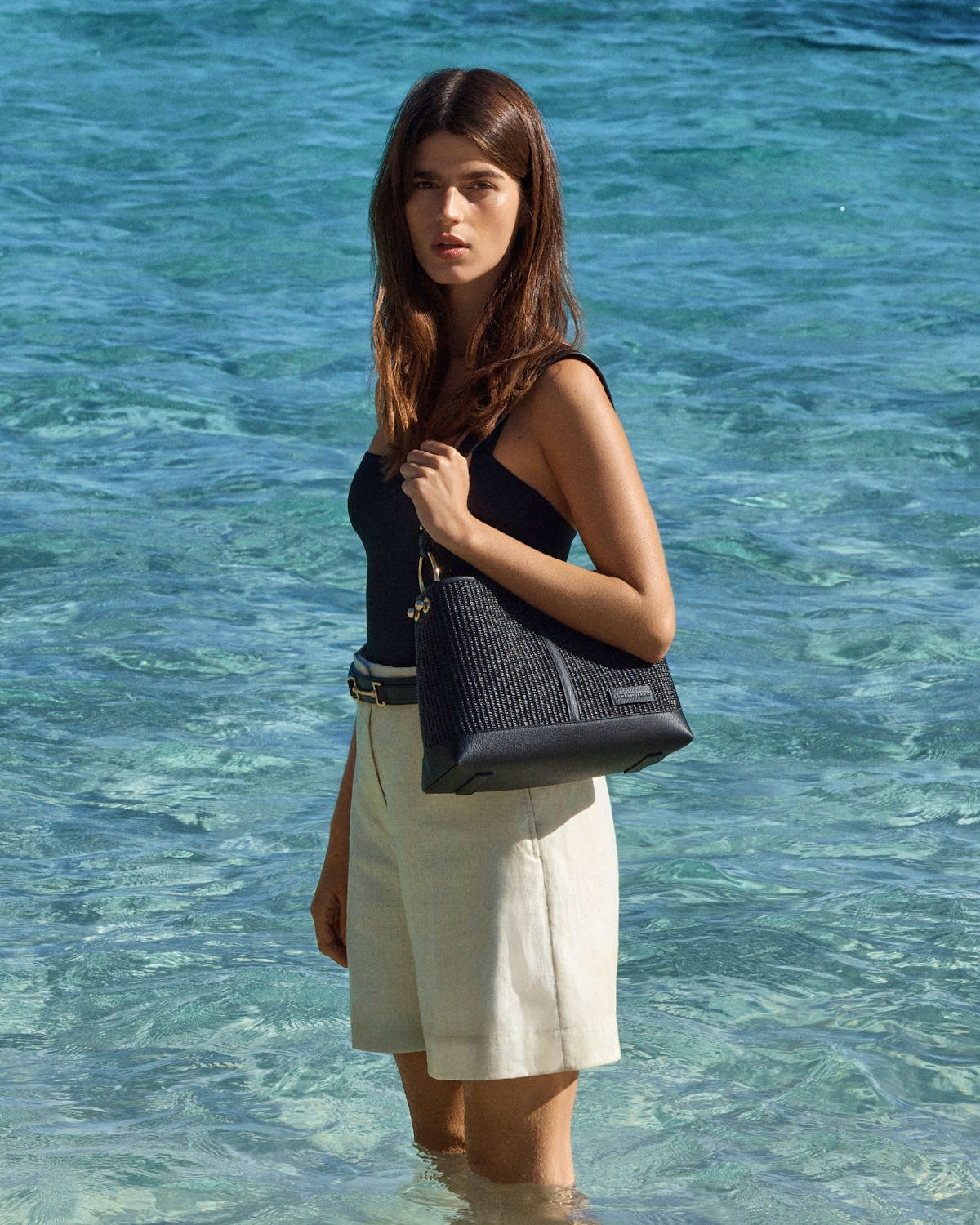 Woman standing in shallow sea holding a black raffia hobo bag.