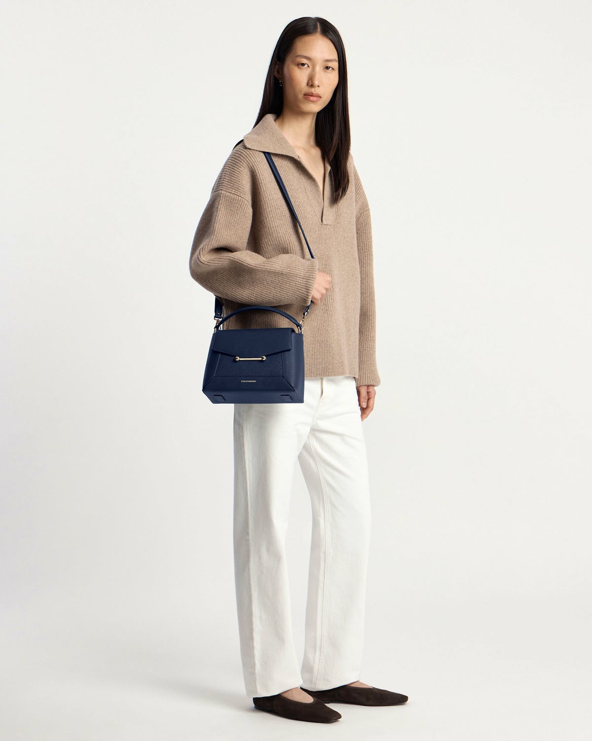 Woman wearing a navy mosaic bag on her shoulder, beige sweater and white trousers.