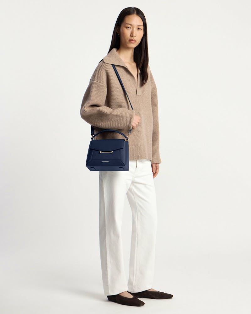 Woman wearing a navy mosaic bag on her shoulder, beige sweater and white trousers.