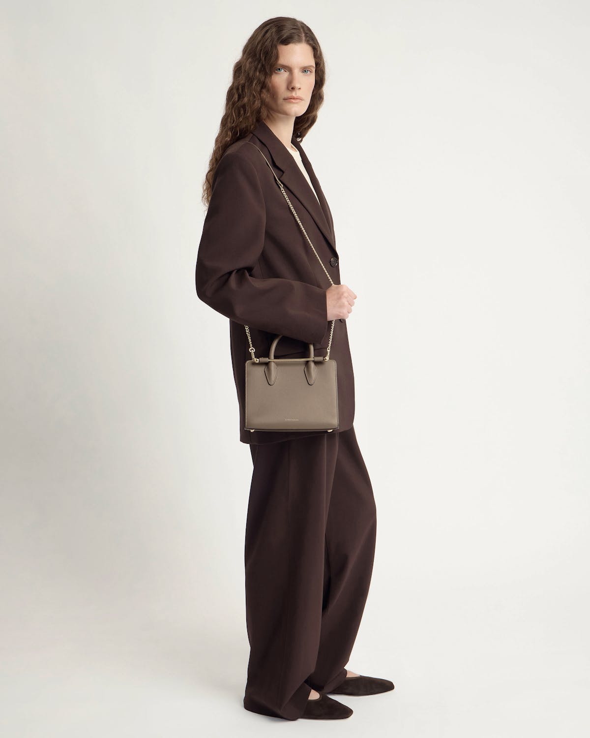Model wearing brown suit carrying taupe mini tote bag with chain strap and top handles.