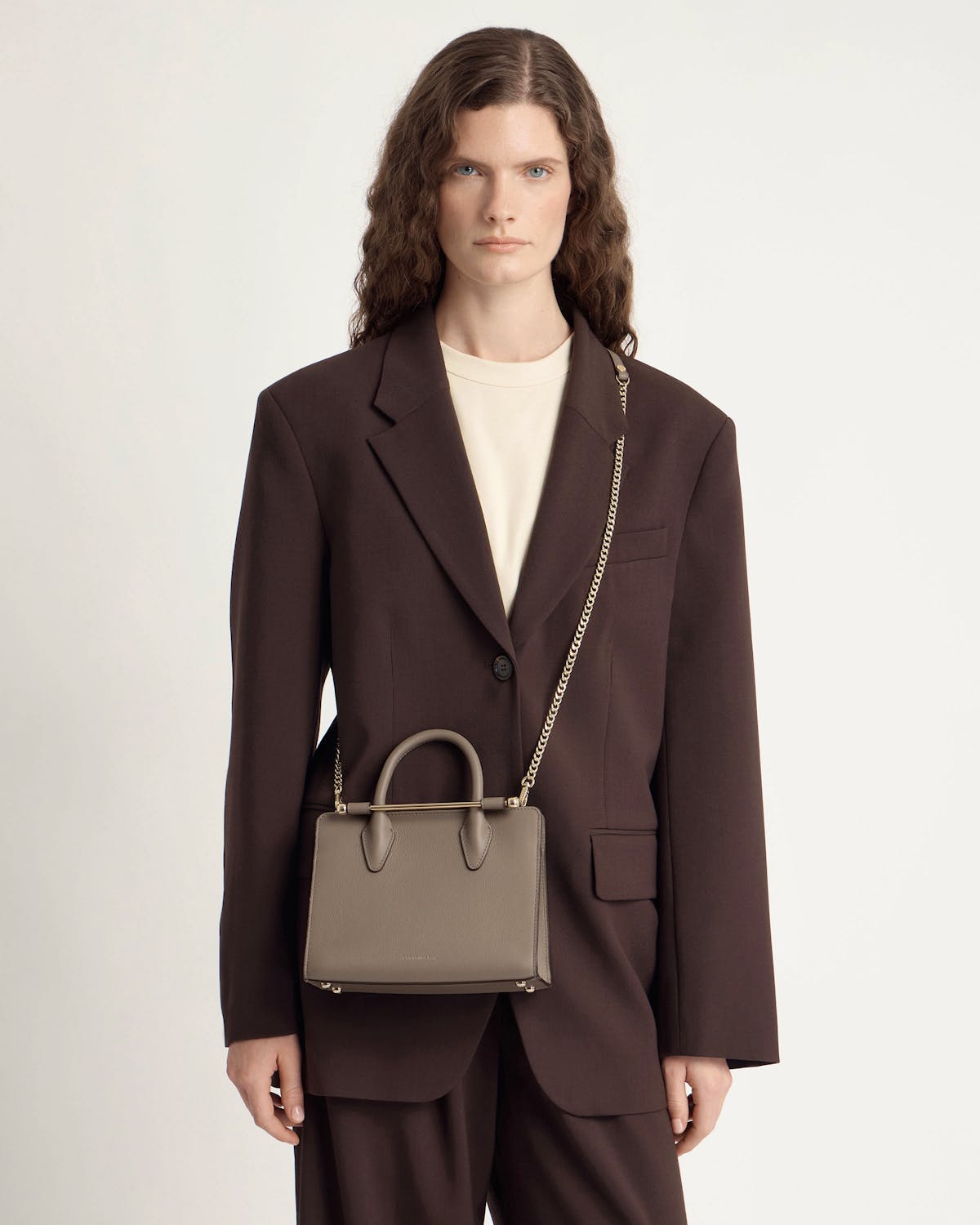 Model wearing a dark brown blazer carrying a taupe mini tote with chain strap.