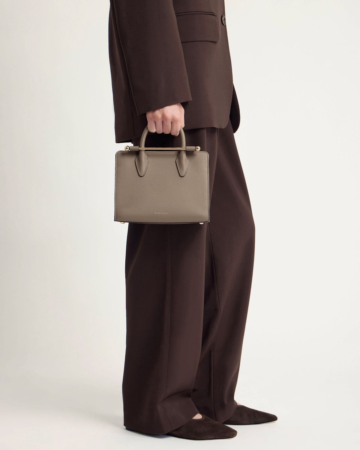 Model holding a taupe mini tote by top handles; metal bar detail, brown suit.
