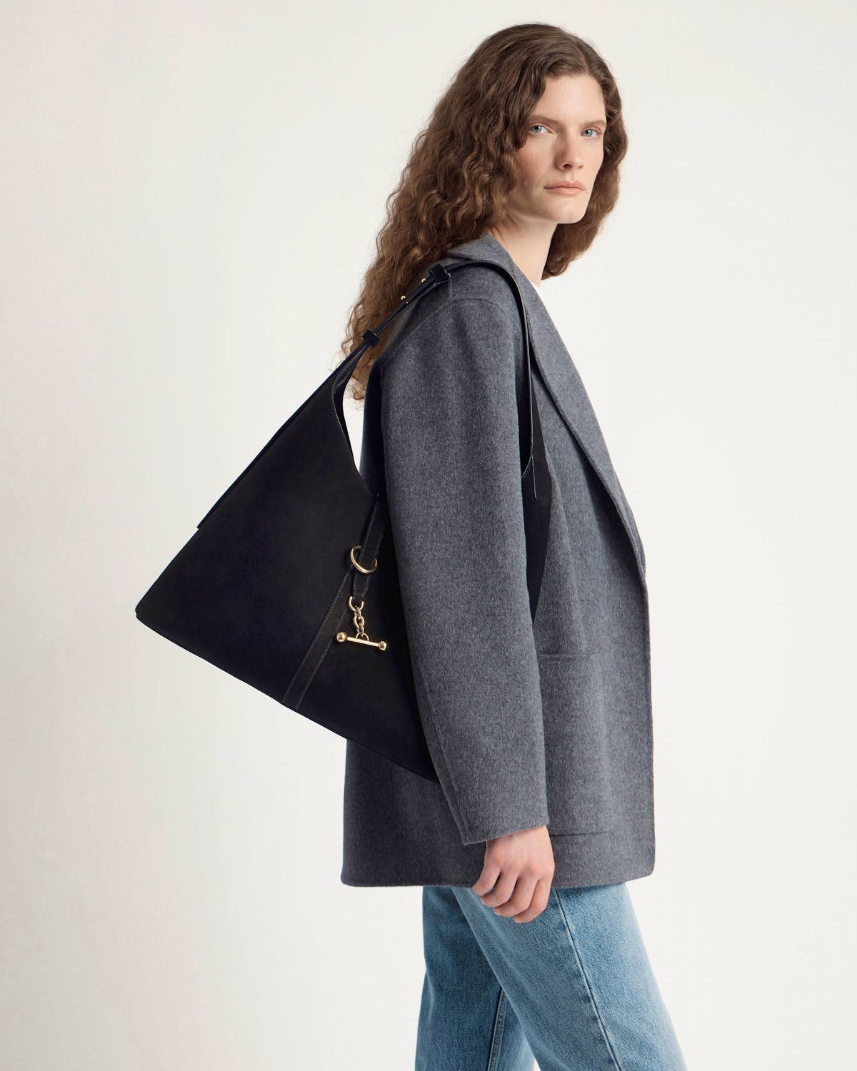 Model wearing grey coat and jeans carrying black suede hobo bag with gold hardware.