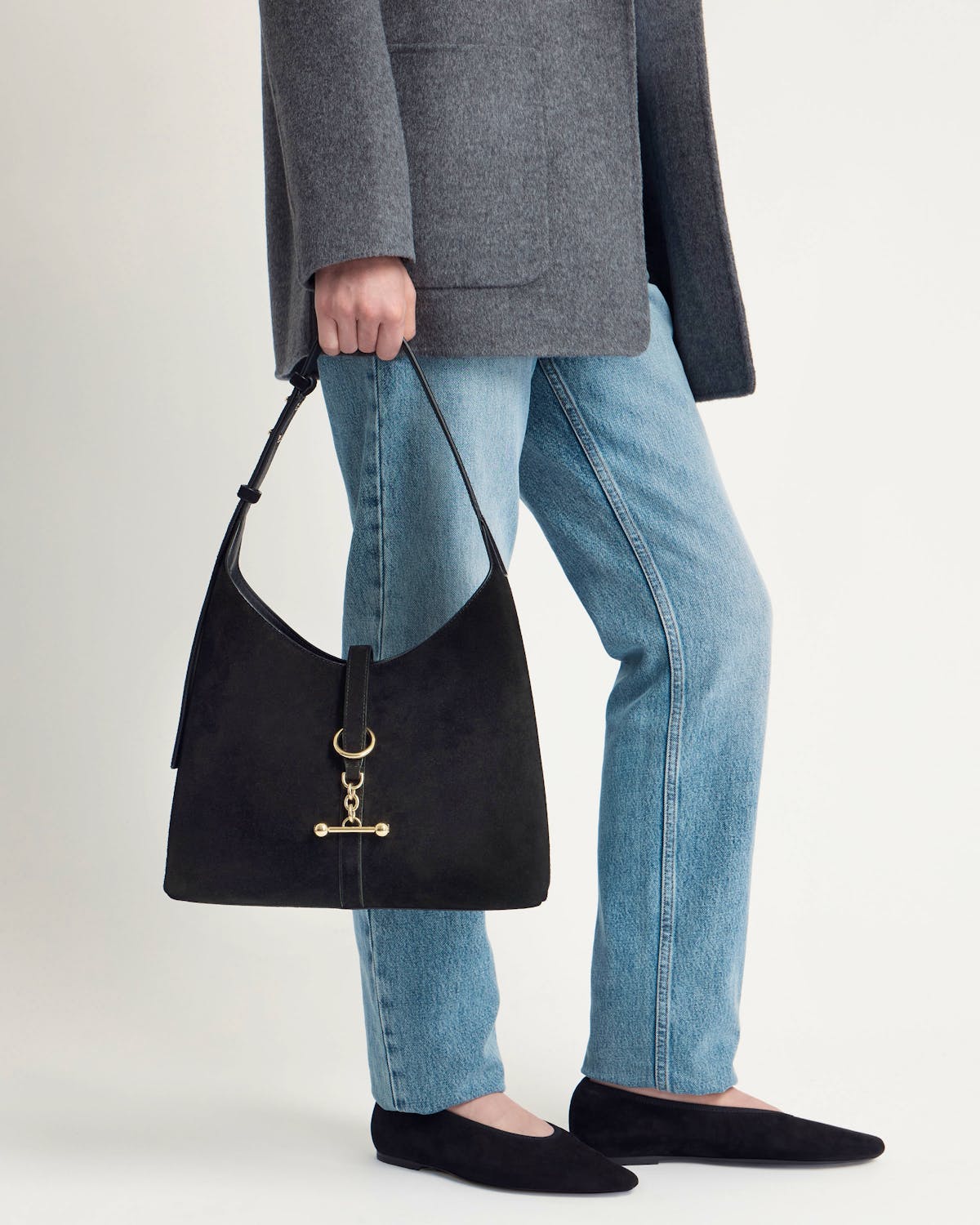 Model holding black suede hobo bag with gold hardware, wearing grey coat and blue jeans