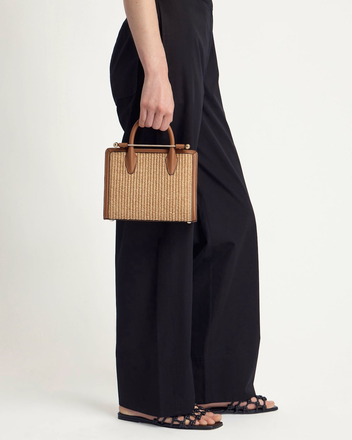 Hand holding tan leather-trimmed mini tote with natural raffia panels, model in black trousers.