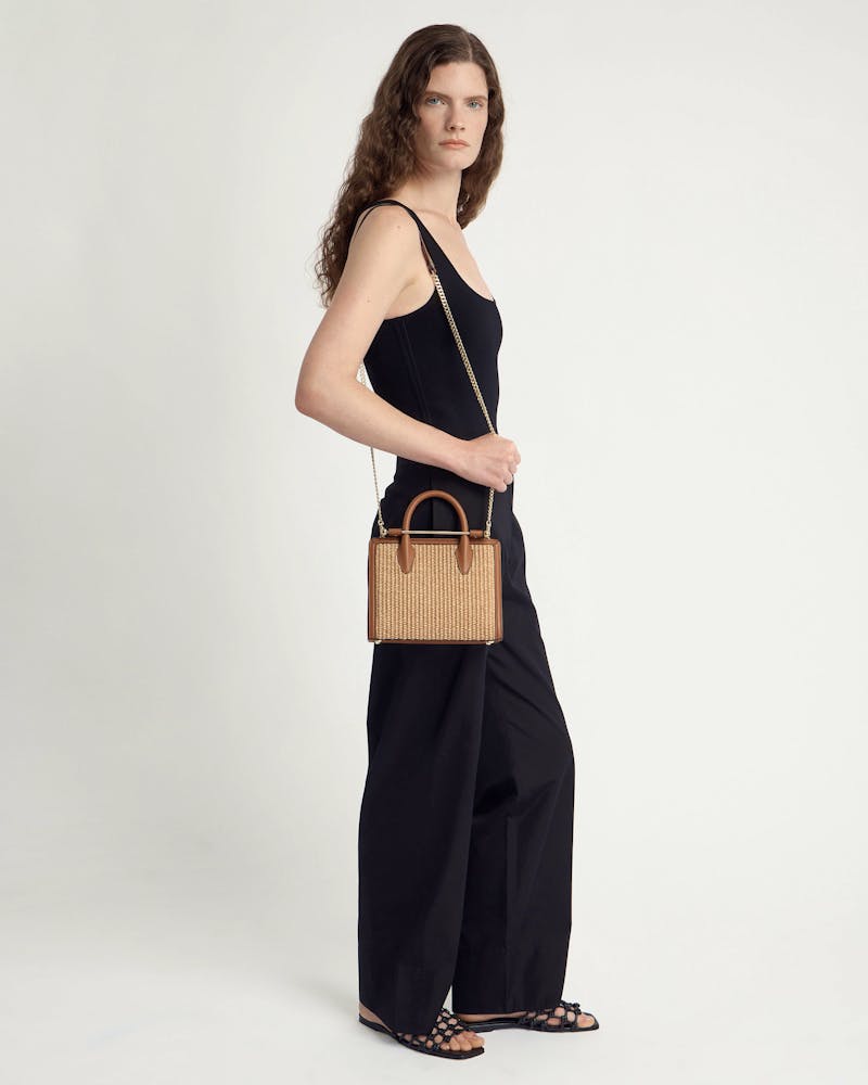 Model wearing black sleeveless outfit holding tan raffia mini tote with gold chain.