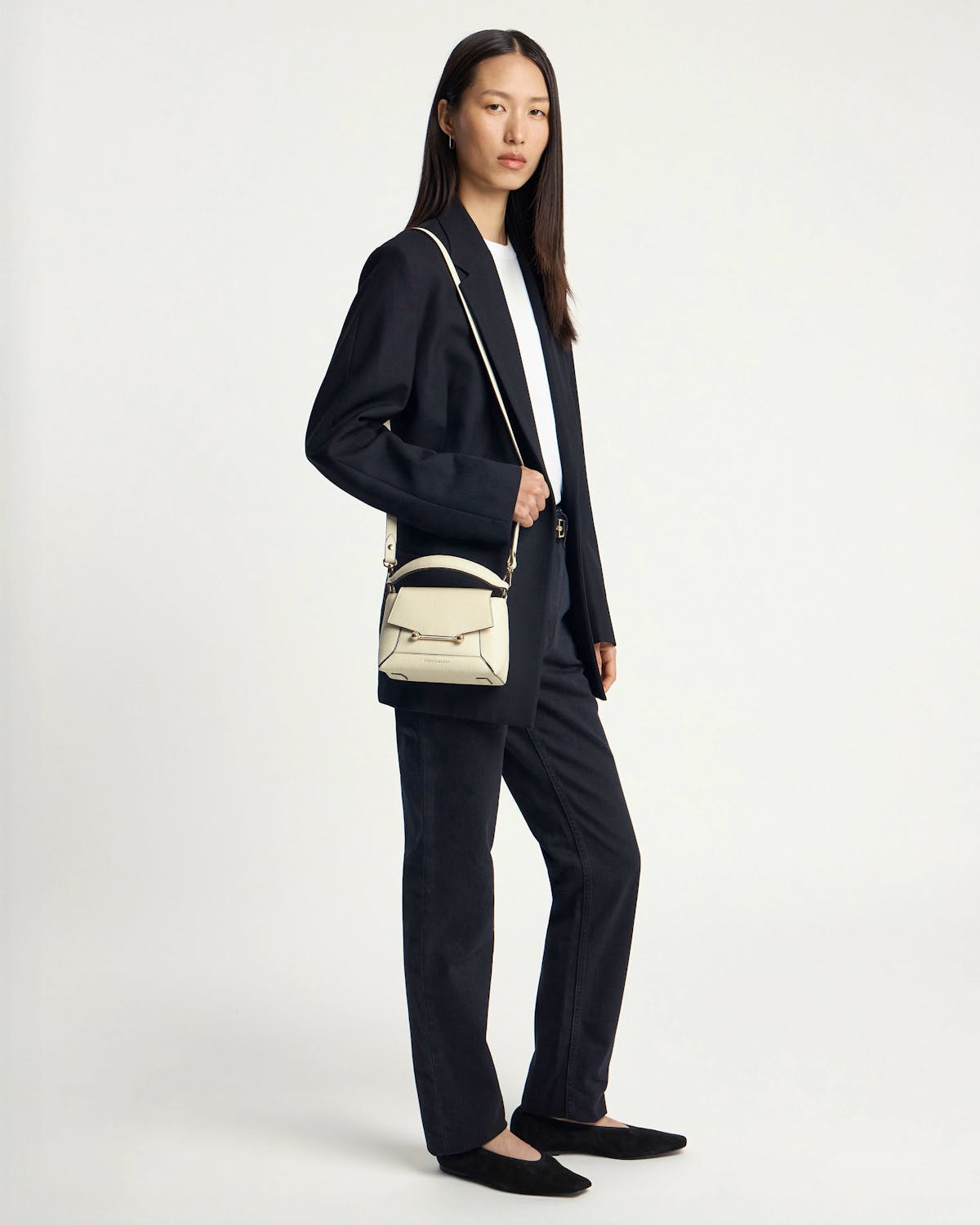 Model wearing black blazer carries small vanilla leather crossbody bag with top handle