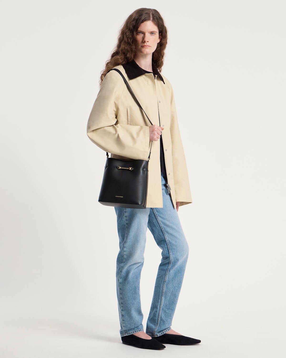Model wearing beige coat and jeans carrying black Corda Bucket bag with Strathberry logo.