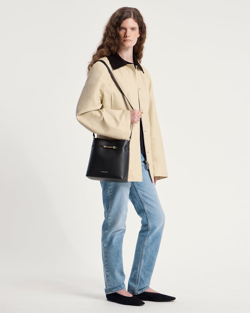 Model wearing beige coat and jeans carrying black Corda Bucket bag with Strathberry logo.