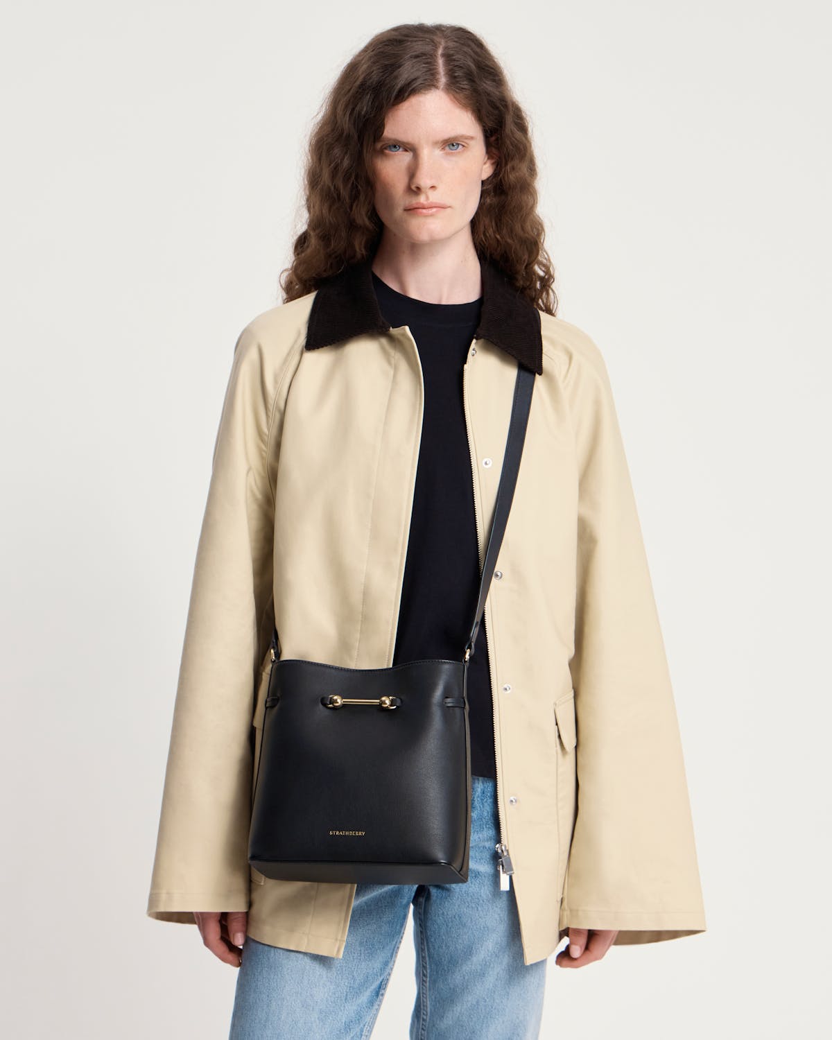 Model wearing beige coat with a black Strathberry Corda bucket bag worn crossbody
