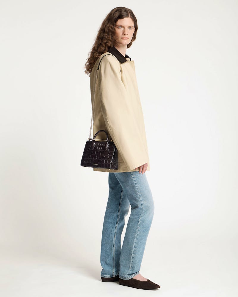 Model wearing beige coat and jeans carrying dark croc mini tote on shoulder.