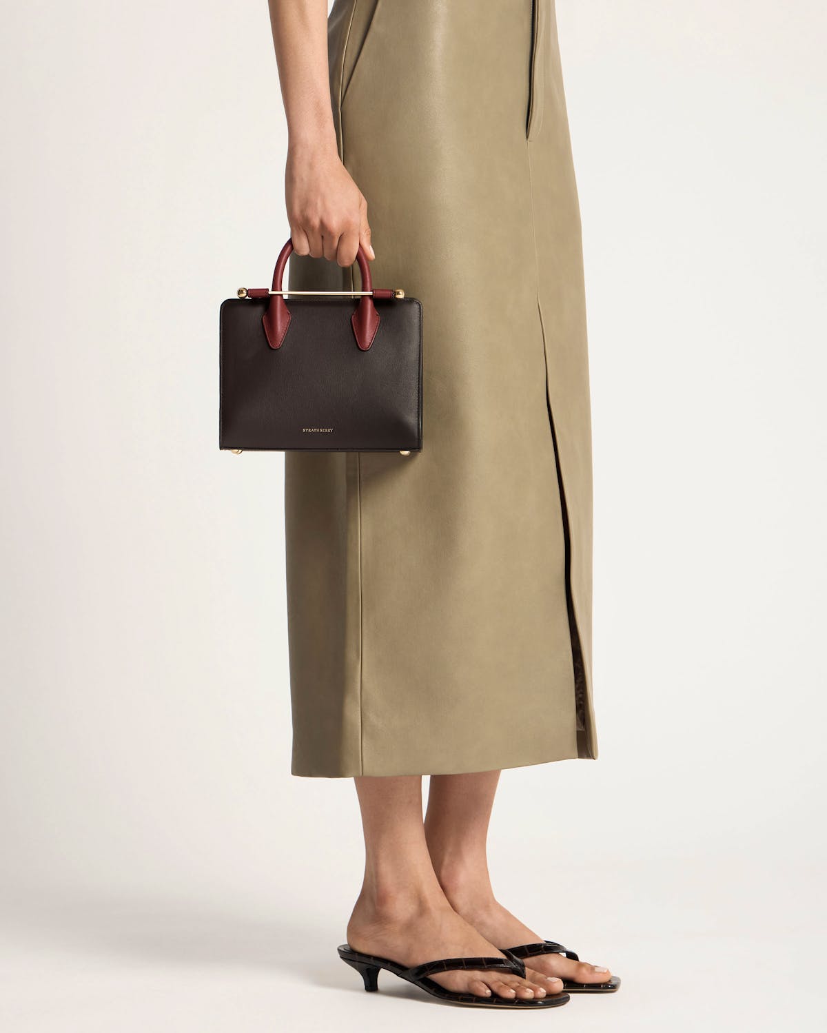 Model's lower body holding Strathberry mini tote in espresso, oxblood handles, black trim