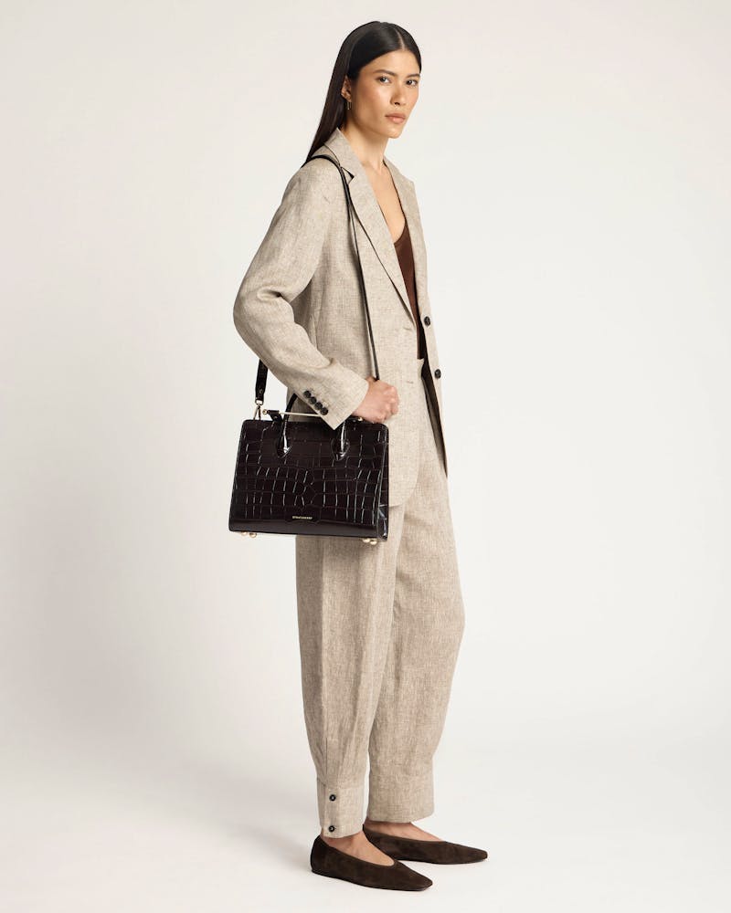 Model in beige suit holding black croc midi tote with Strathberry logo.