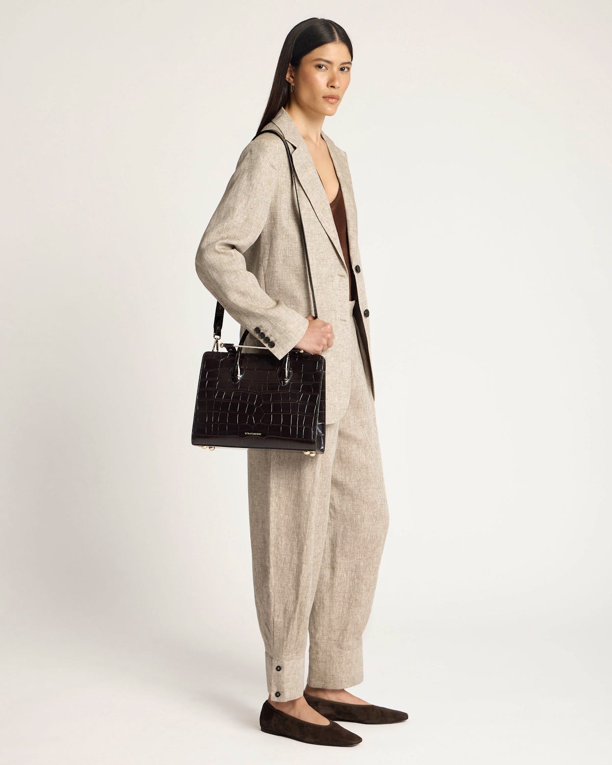 Model in beige suit holding black croc midi tote with Strathberry logo.