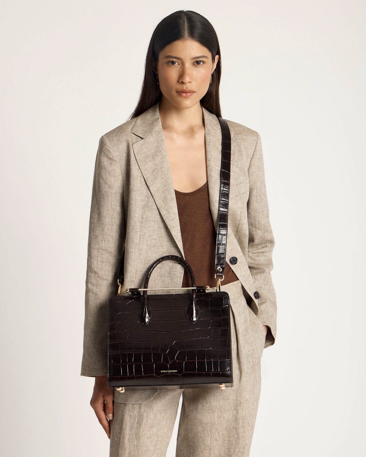 Woman in beige blazer holding dark croc-embossed midi tote with Strathberry logo.