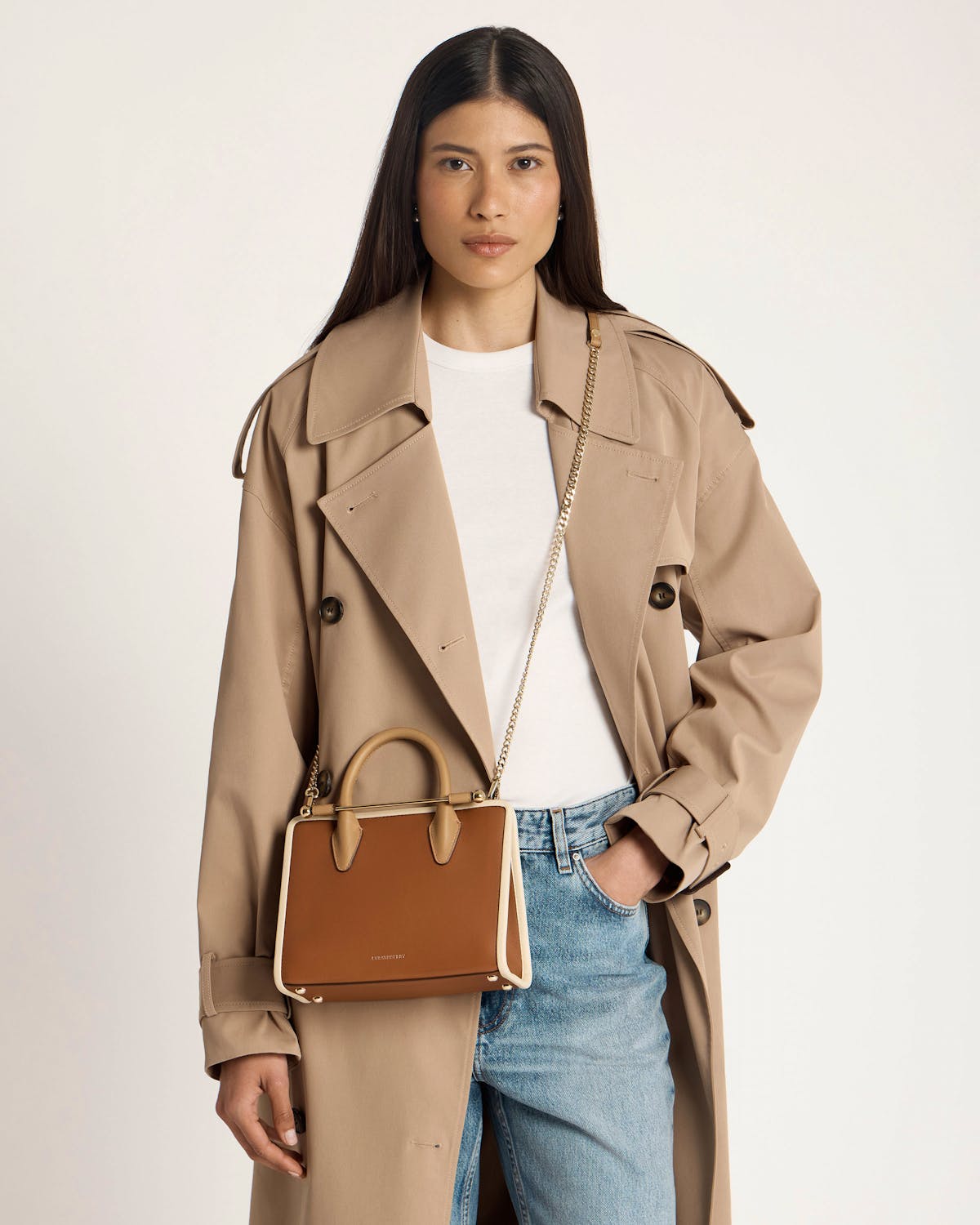 Model wearing a beige trench and white tee with a Strathberry caramel mini tote.