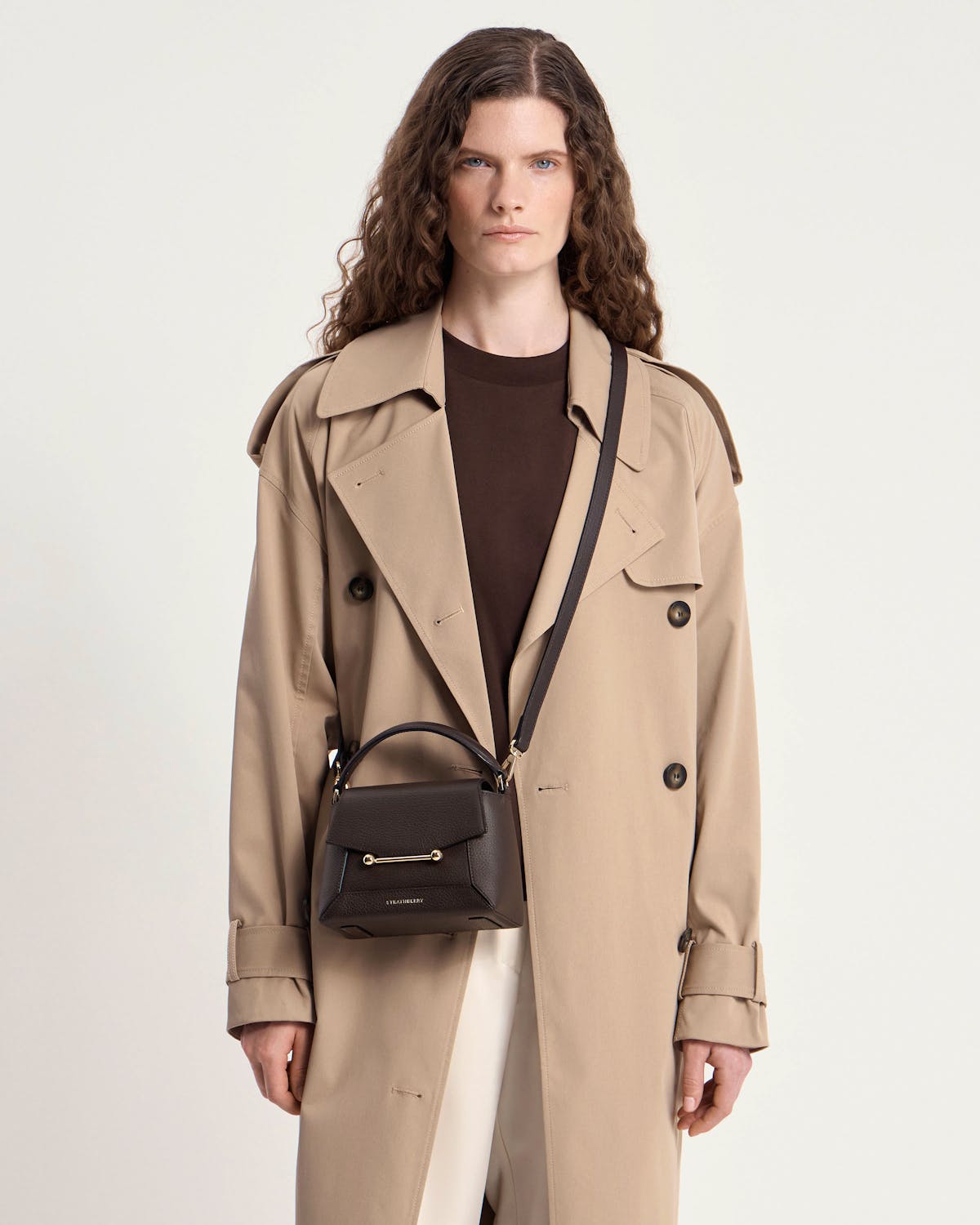 Model wearing trench coat with small Strathberry Mosaic Nano espresso leather top-handle bag.