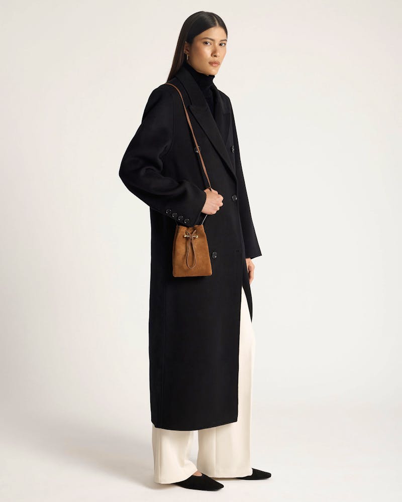 Model wearing black coat and roll-neck, carrying a tan suede drawstring midi pouch.