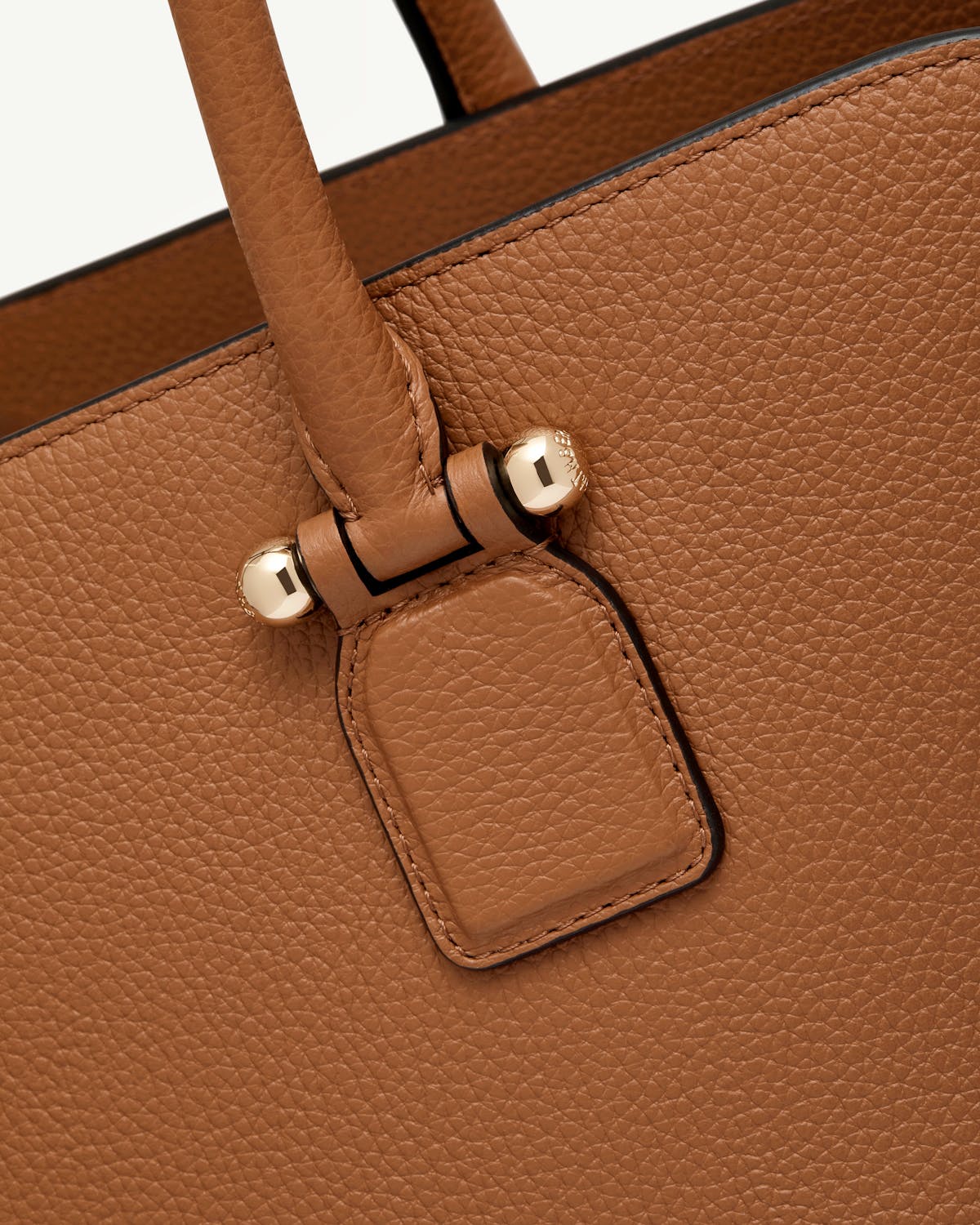 Tan pebbled leather bag showing handle attachment with gold studs.