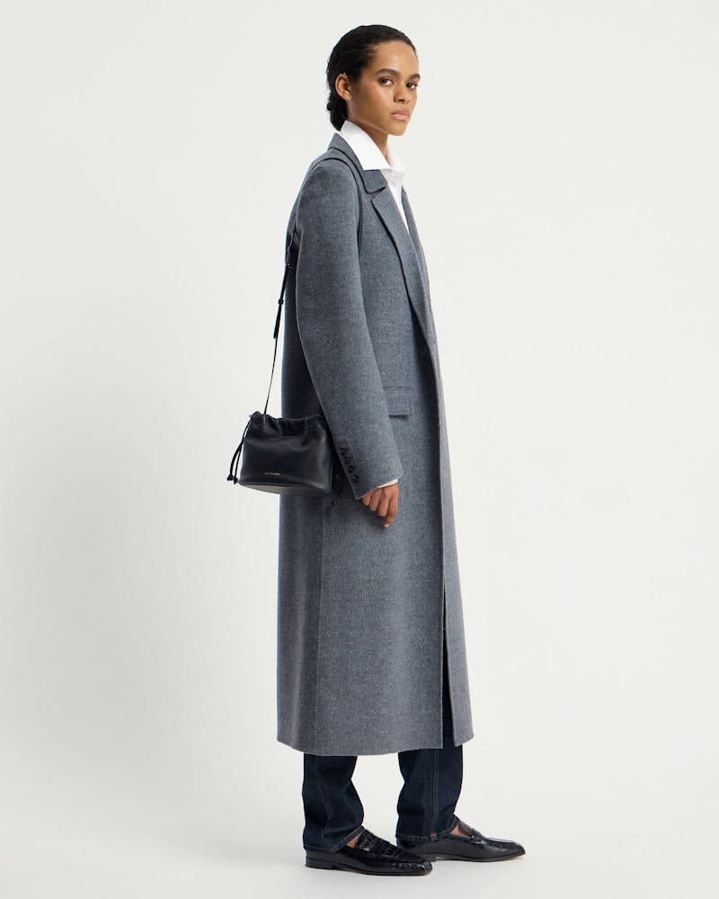 Model in grey coat carrying a black drawstring shoulder bag at her side.