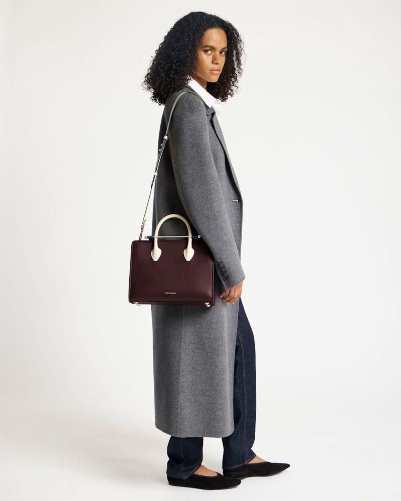 Model wearing a grey coat holding a Strathberry midi tote in burgundy, navy and vanilla