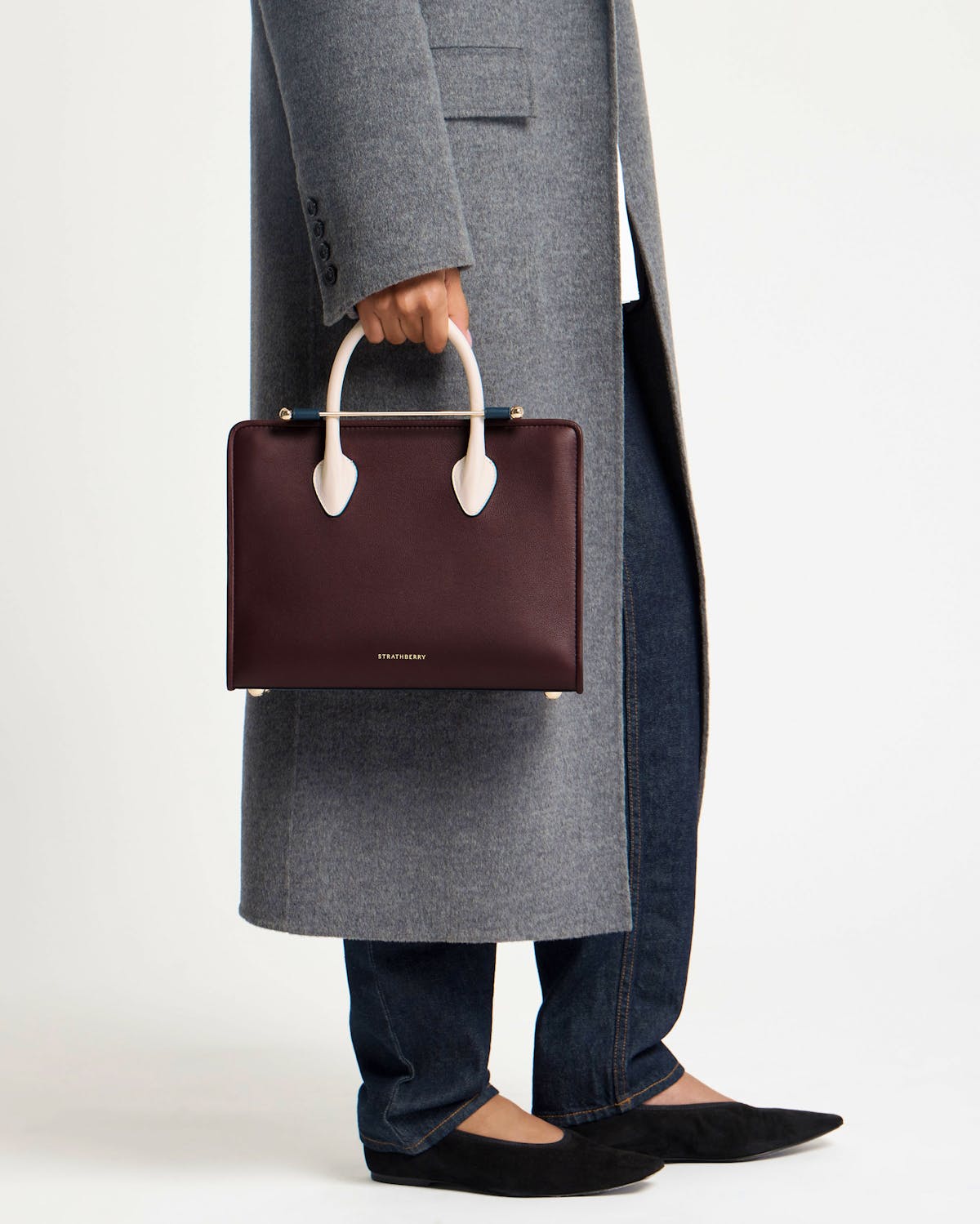 Model cropped below waist holding Strathberry midi tote in burgundy with vanilla and navy handles.