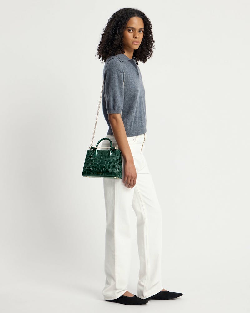 Model wearing grey top and white trousers with green croc mini tote at her side.