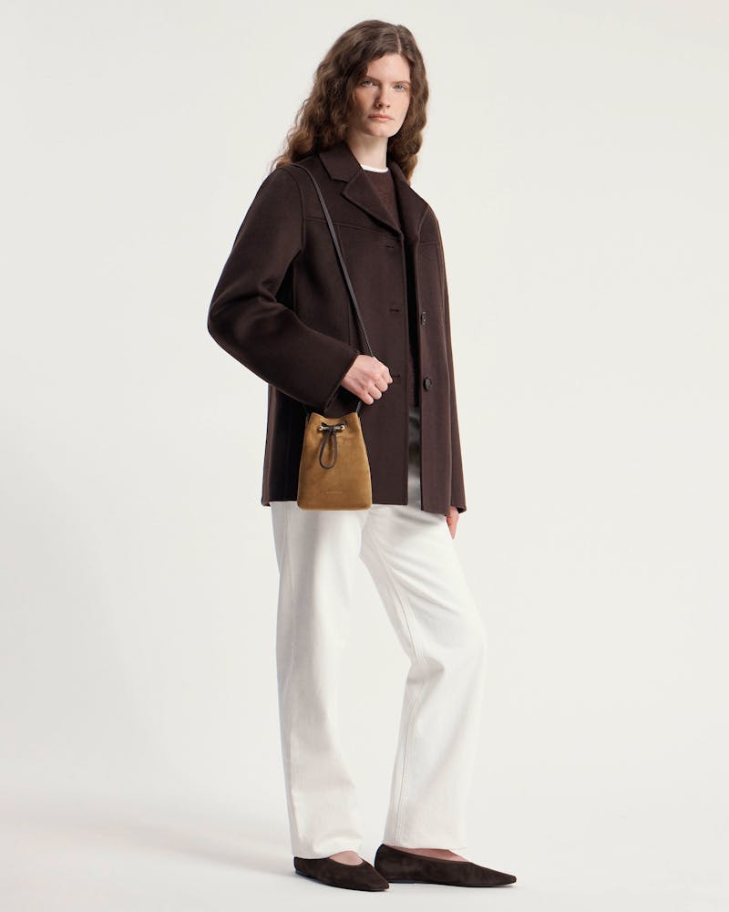 Woman in dark brown coat and white trousers carrying toffee suede drawstring pouch.