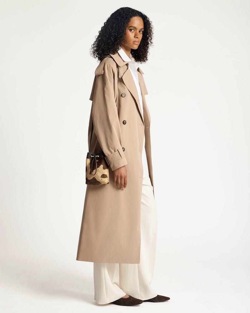Woman in beige trench coat holding a sand-and-espresso spot-print midi pouch.