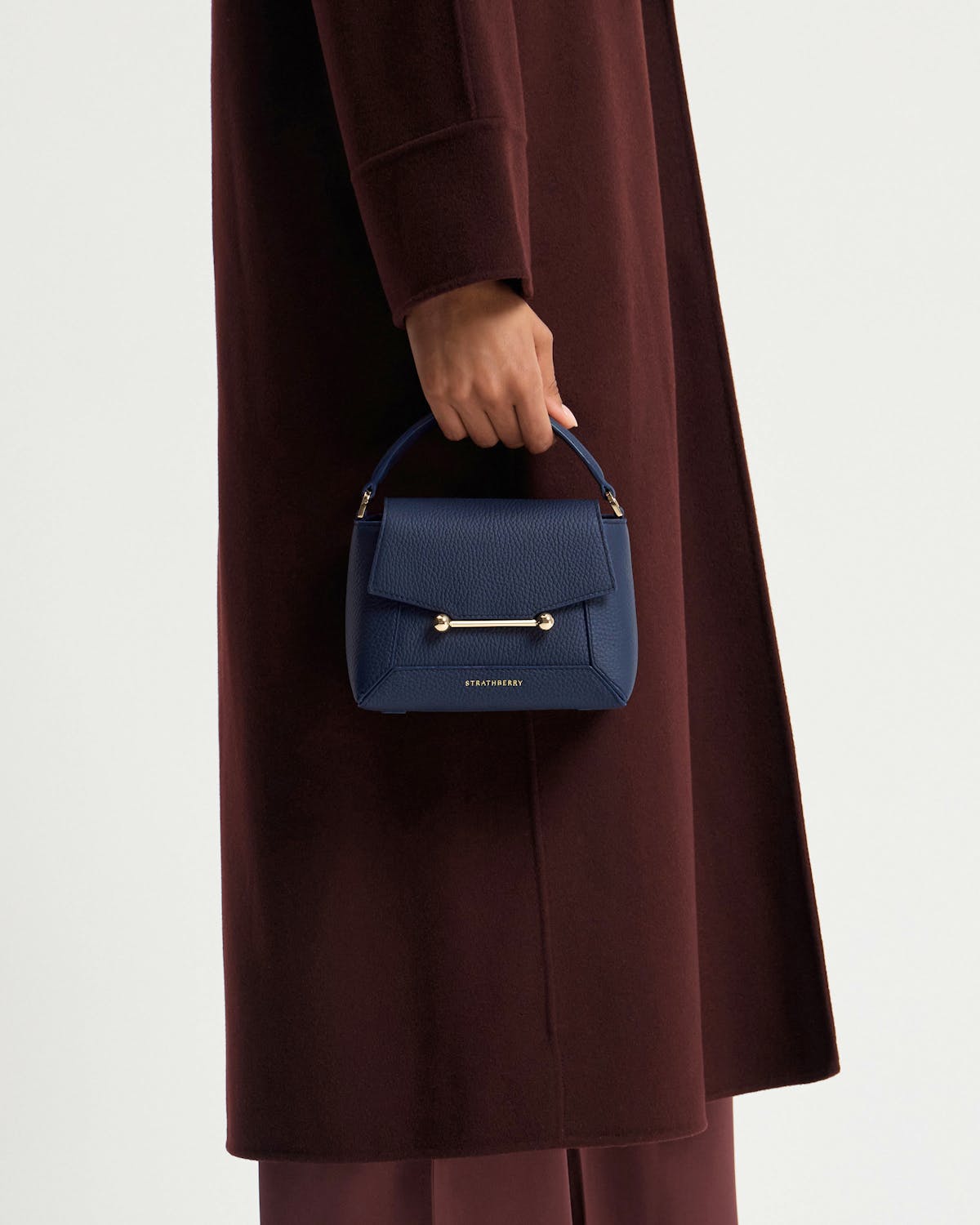 Model's hand holding navy Strathberry Mosaic Nano bag against coat.