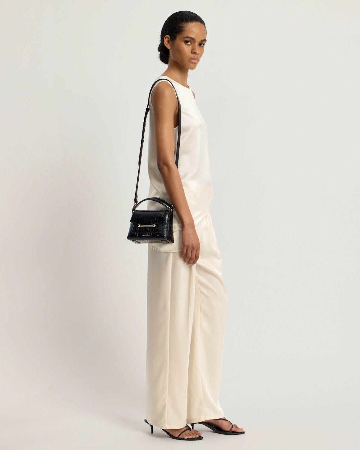 Model wearing cream outfit carrying small black croc-embossed top-handle bag with shoulder strap.