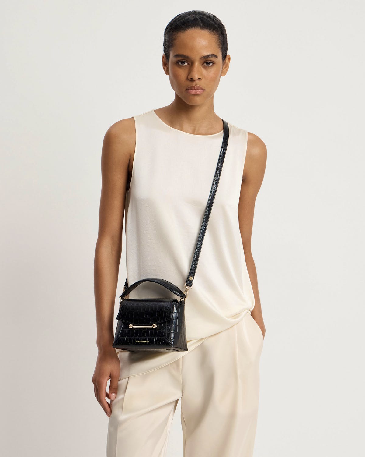Model wearing cream top and small black croc top-handle crossbody bag with Strathberry logo