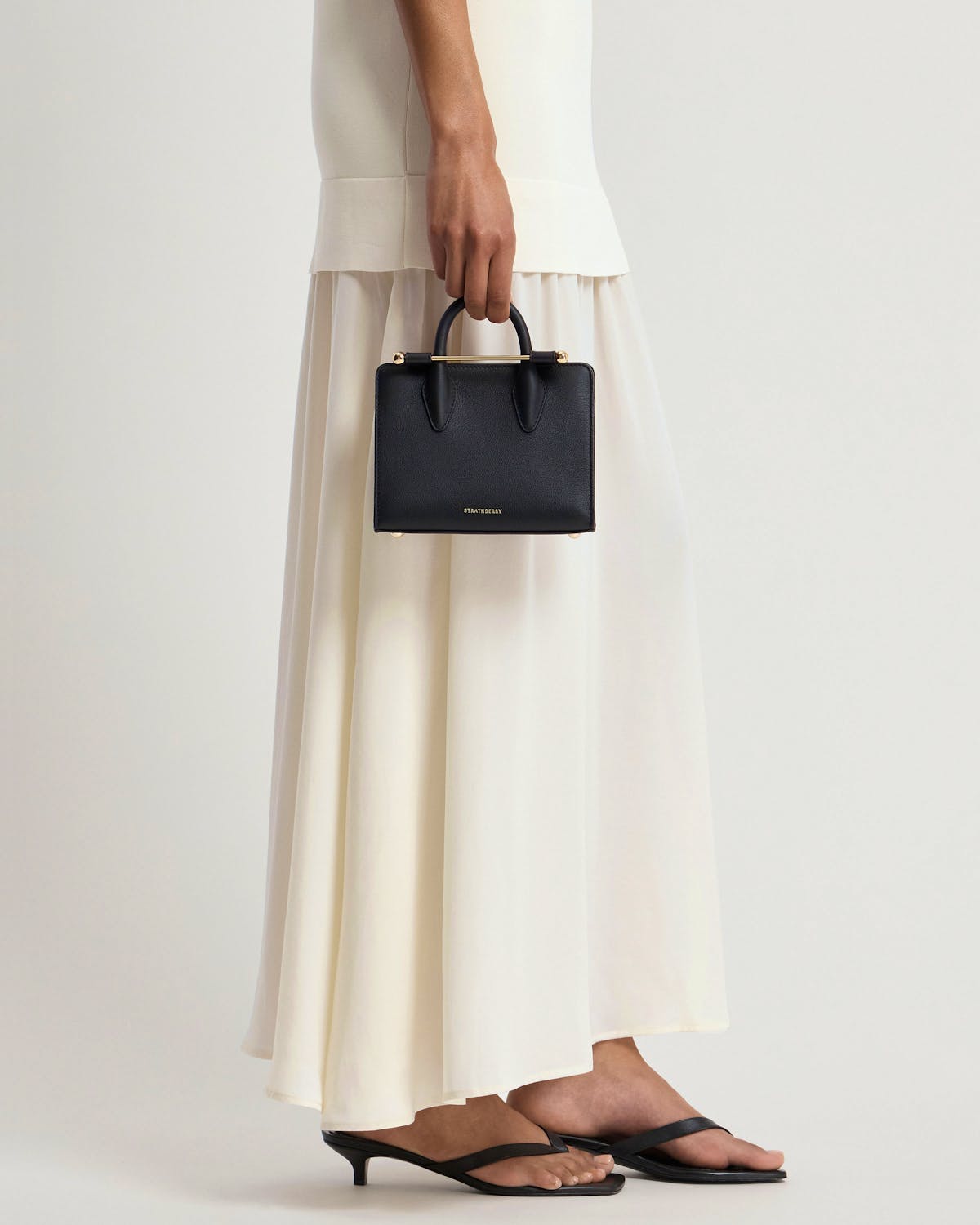 Model holding black Strathberry nano tote with cream-coloured skirt and black sandals.