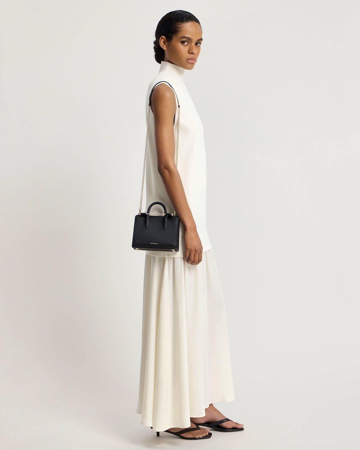 Model wearing white dress carrying black Strathberry nano tote with chain strap.