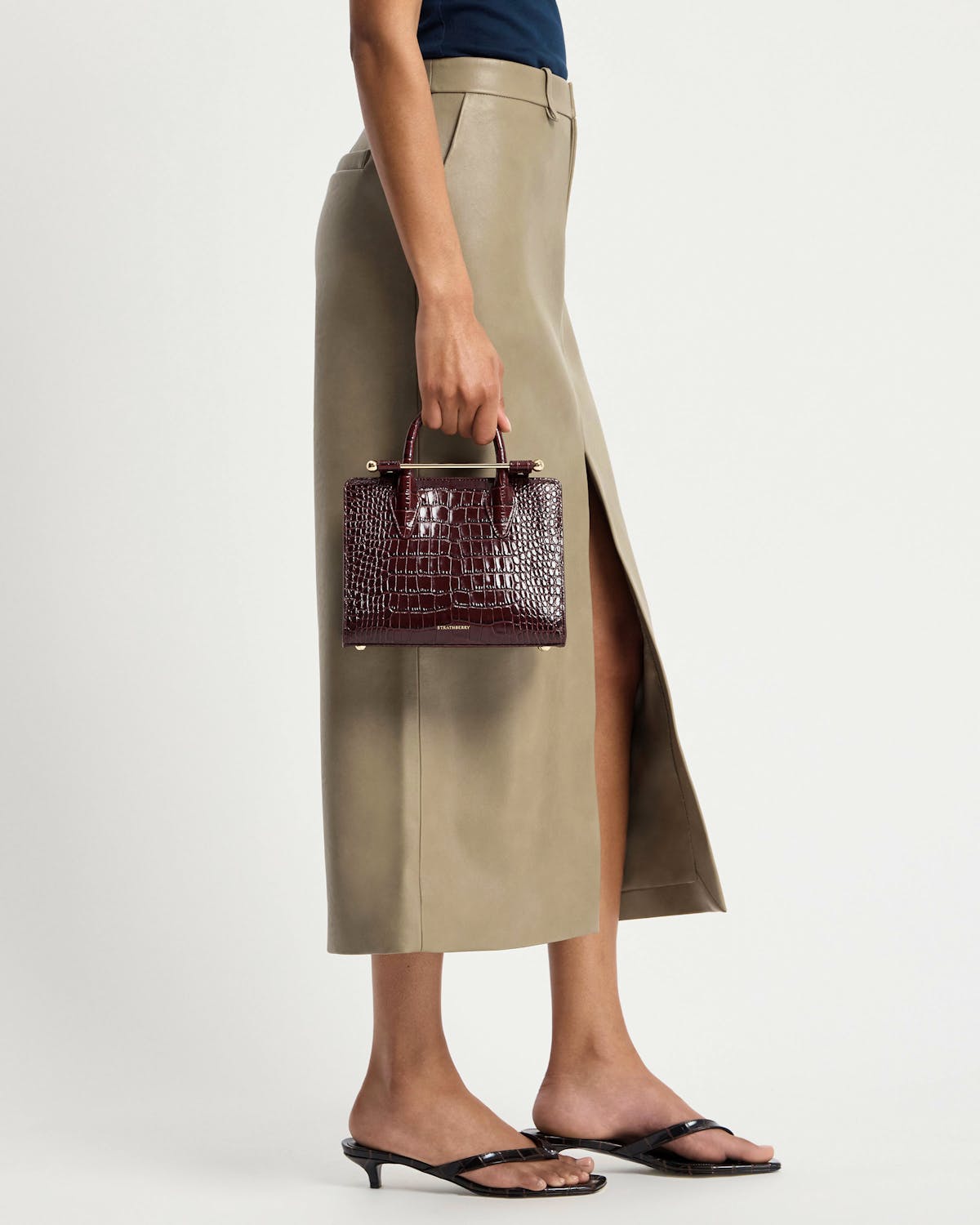 Lower body wearing beige skirt with side slit holding Strathberry burgundy croc mini tote