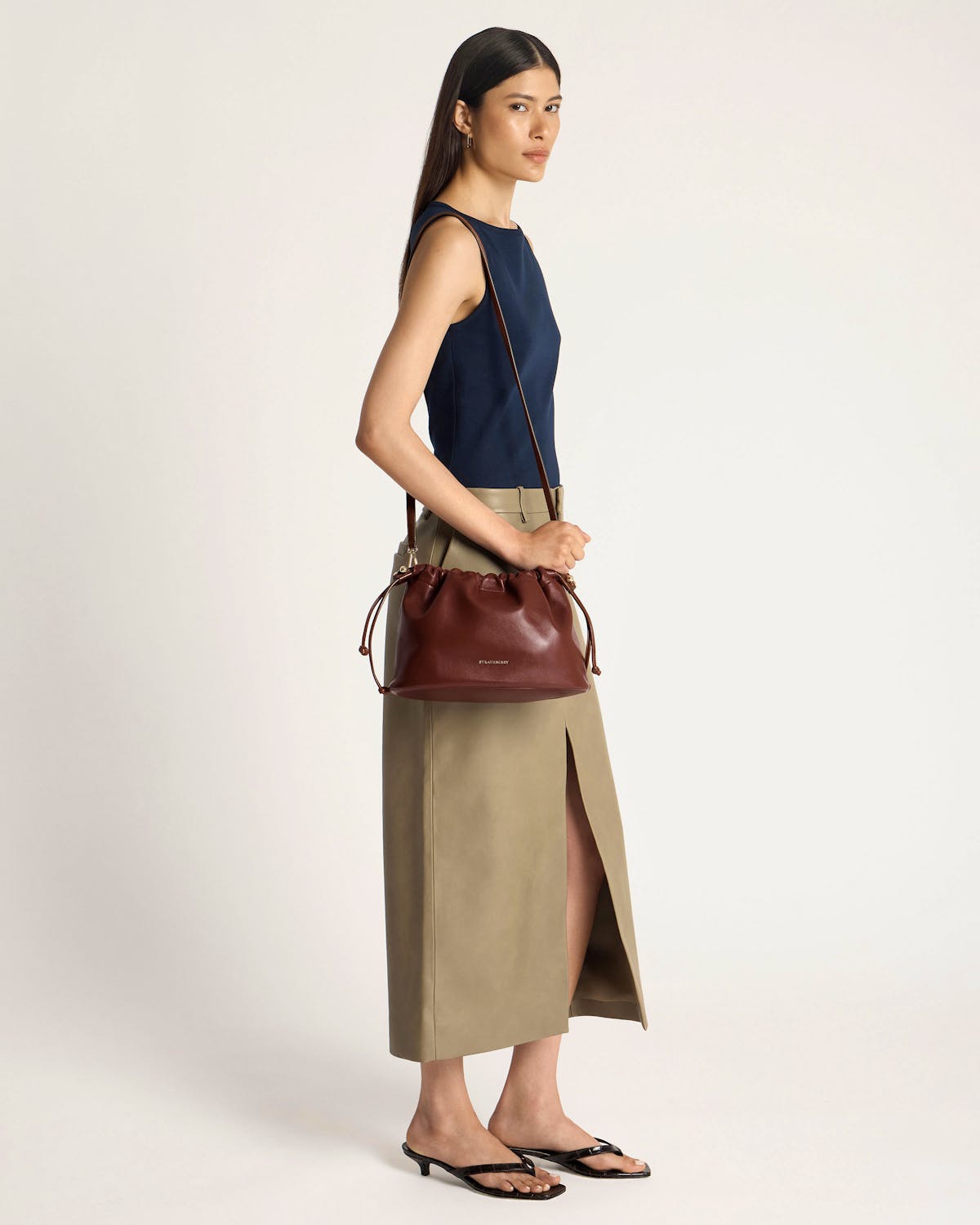 Model wearing navy top and khaki skirt holding walnut Charlotte midi drawstring bag by Strathberry