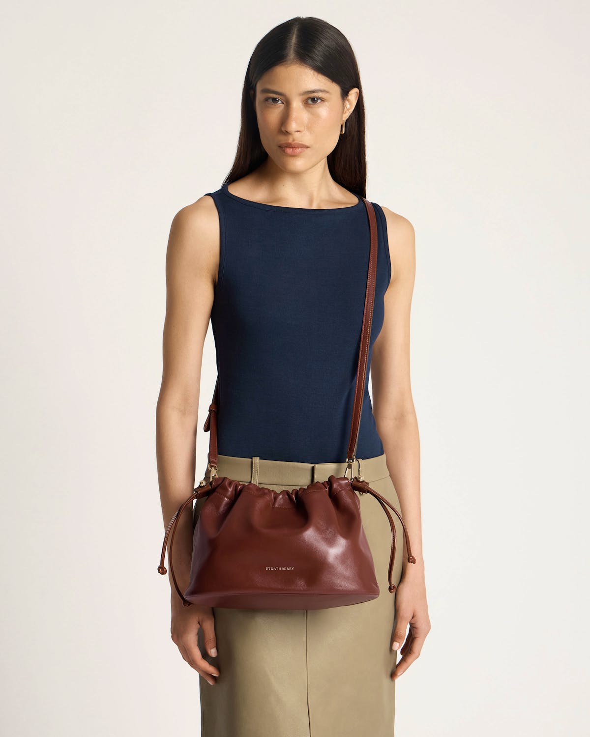 Model in navy top wearing Strathberry Charlotte Midi drawstring bag in walnut on shoulder.