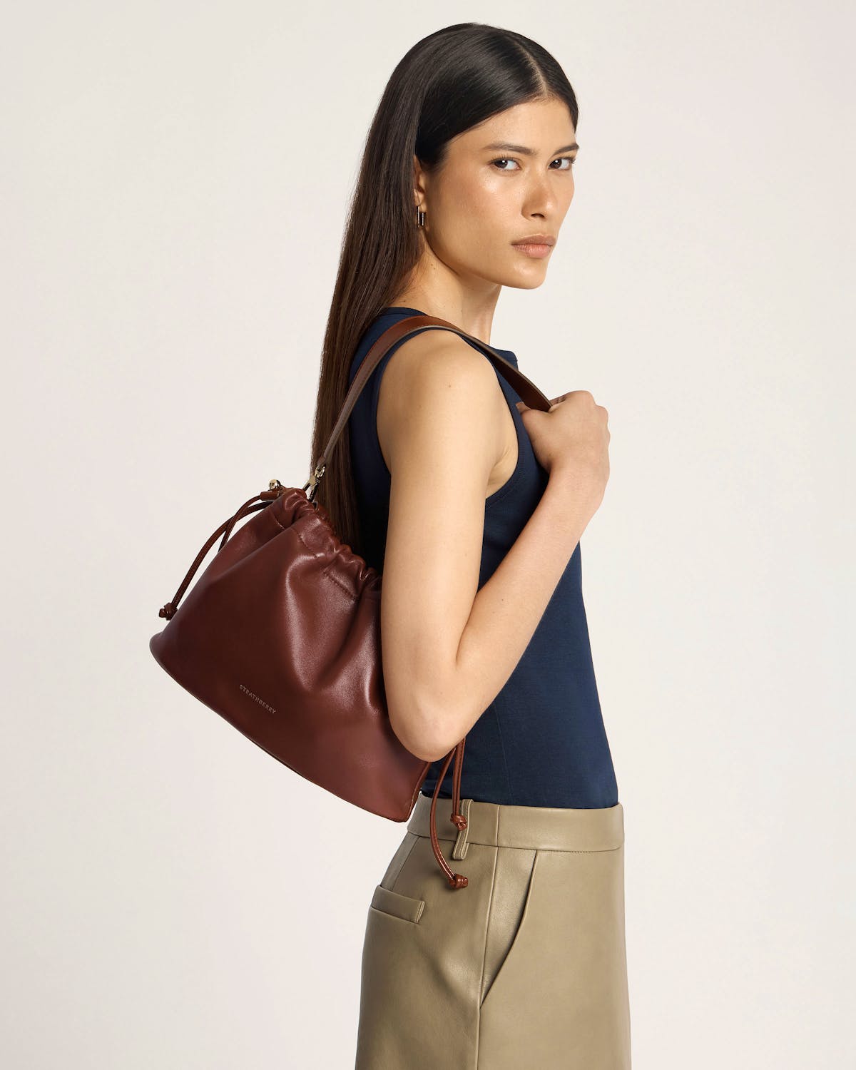 Model wearing navy top carrying walnut Strathberry Charlotte Midi Drawstring bag on shoulder.