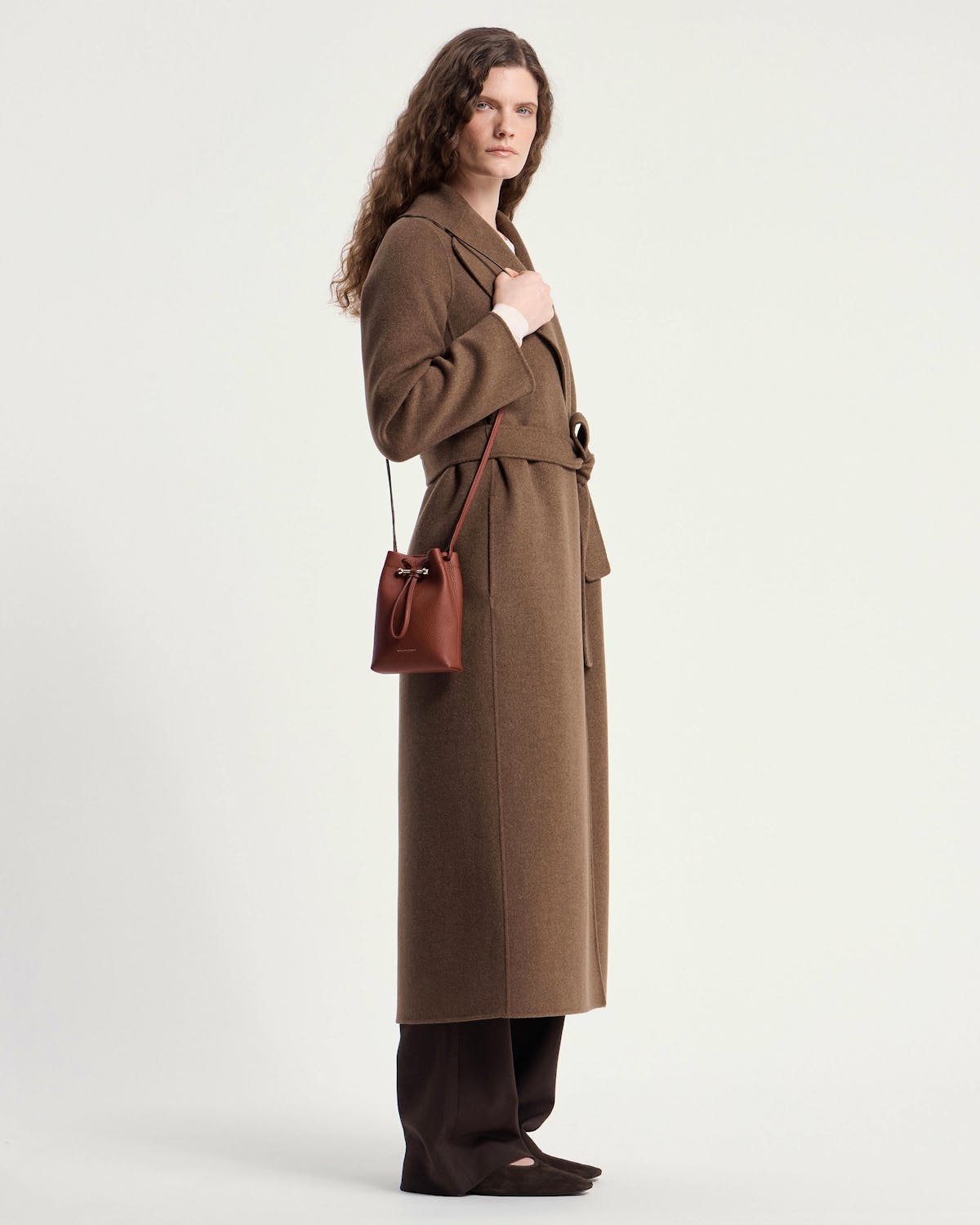 Model in brown coat carrying a walnut leather midi pouch on shoulder.