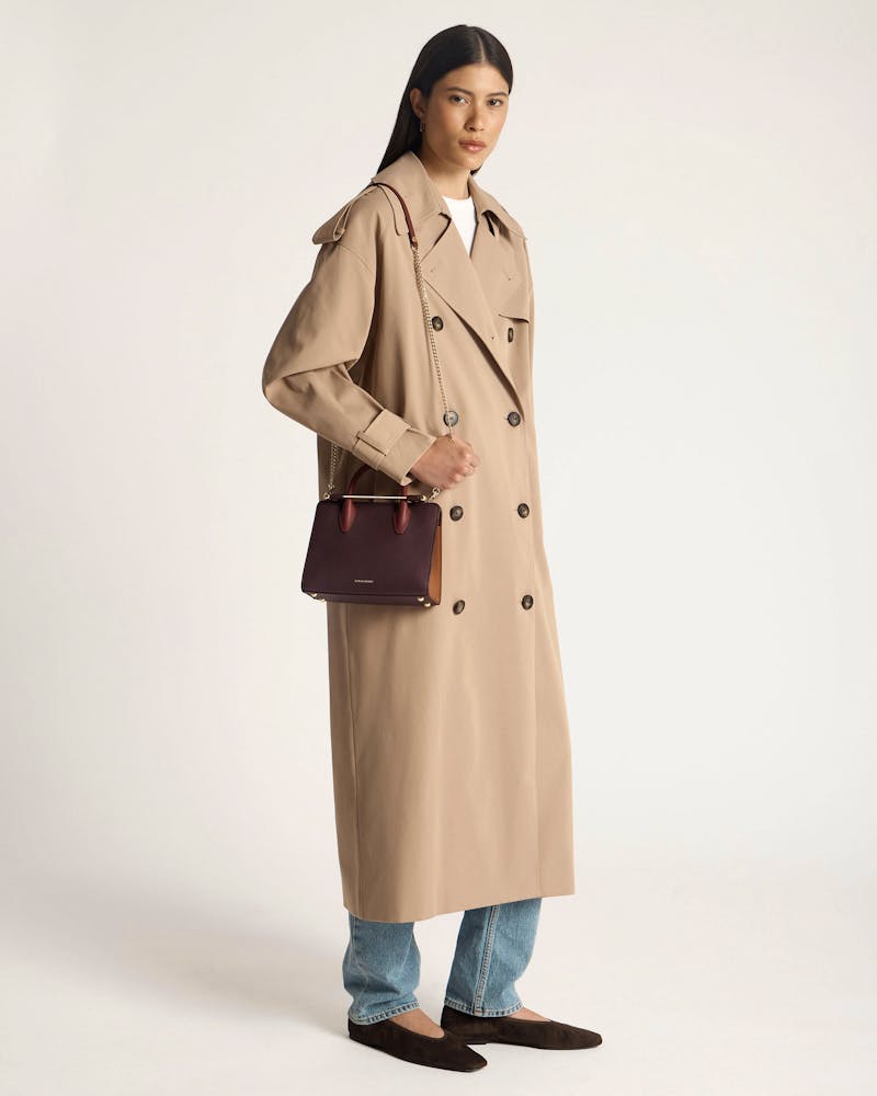 Model in beige trench coat holding burgundy, chestnut and walnut Strathberry mini tote bag
