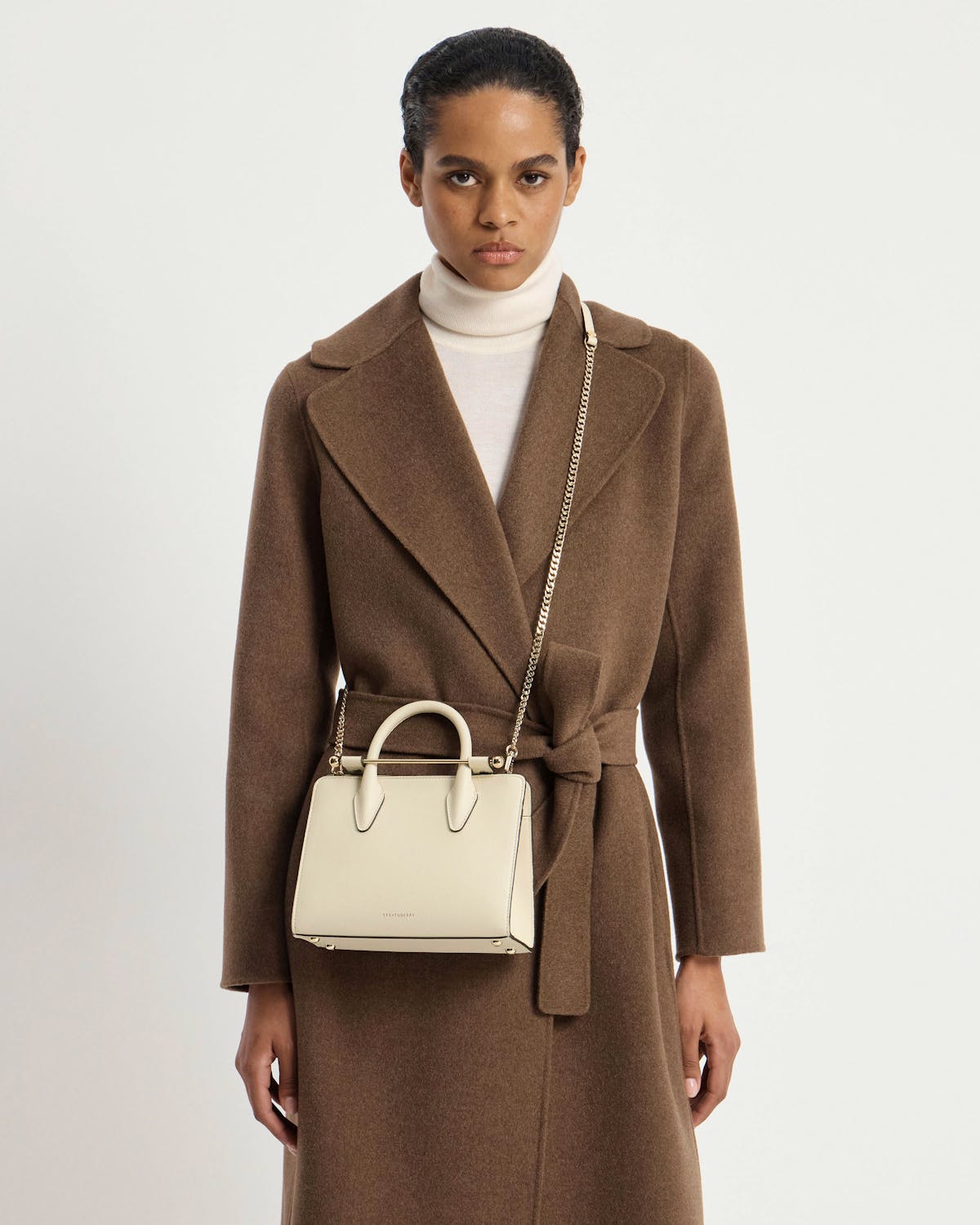 Model wearing brown coat with vanilla Strathberry mini tote on gold chain.