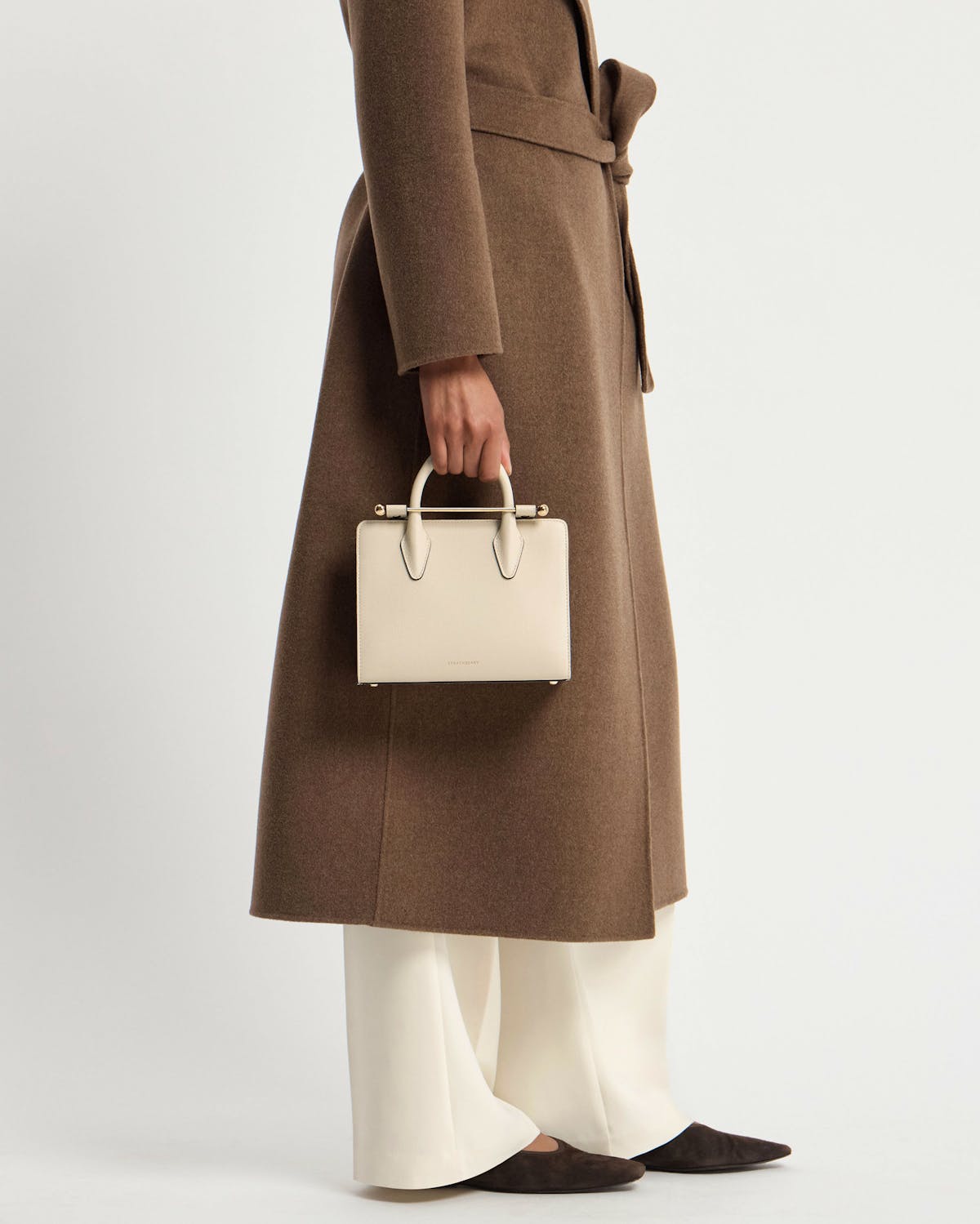 Model cropped below waist wearing brown coat, holding vanilla mini tote by top handles.