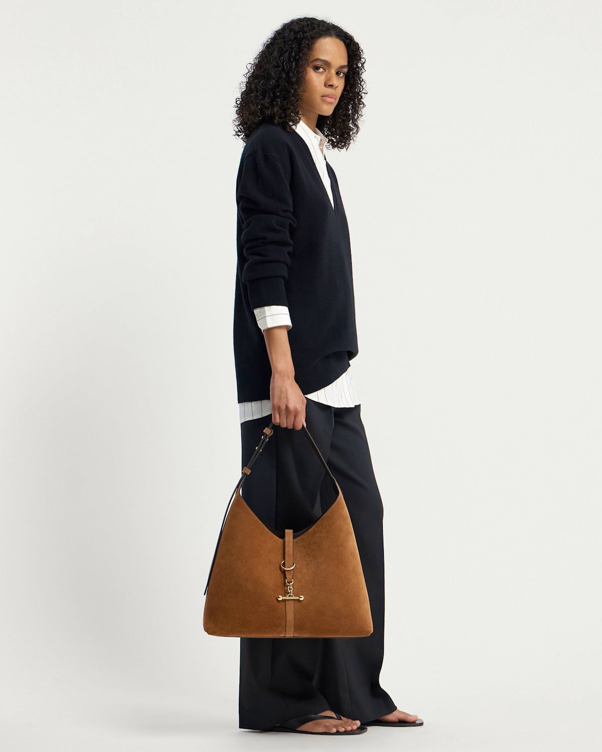 Model holding tan suede hobo bag with gold hardware, wearing black jumper.