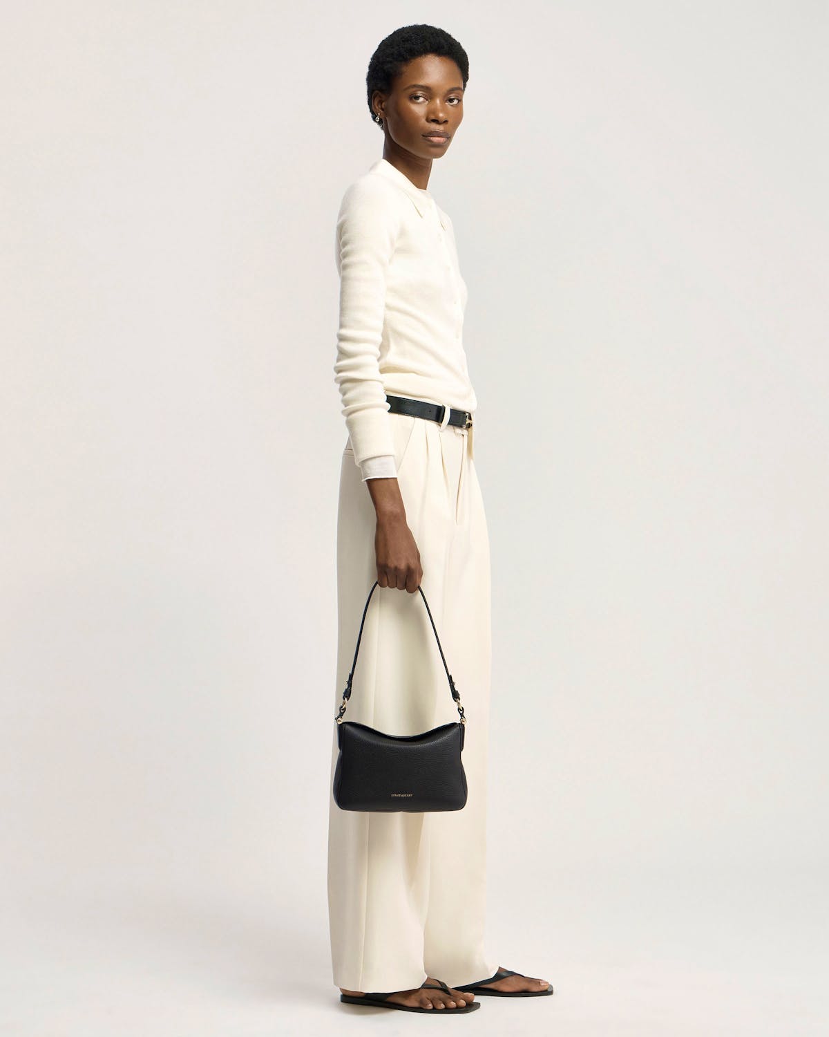 Model holding black Sway shoulder bag by Strathberry, wearing white top and cream trousers.